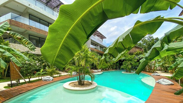 Una piscina con cubierta de madera, rodeada de plantas tropicales y un edificio moderno al fondo.