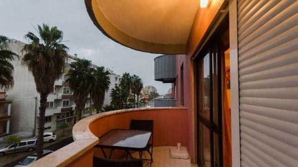 A balcony with a table and chairs, overlooking a street with palm trees and buildings. - Next to Lisbon 100m from Beaches & Surf (Costa da Caparica)