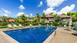 A rectangular swimming pool with clear blue water, surrounded by lounge chairs and umbrellas, set against a backdrop of tropical vegetation and a clear sky. - Tropical Getaway North Shore Princeville Studio (Princeville)
