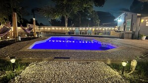 En swimmingpool omgiven av en gång av småsten, med ett trädäck och ljusslingor i bakgrunden.
