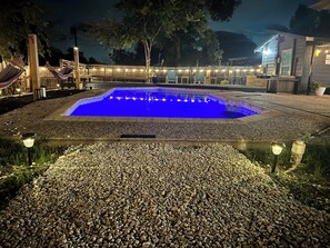 A swimming pool surrounded by a pebble path, with a wooden deck and string lights in the background. - CentralSA 4-bed 3-full bath house w/private heated-pool (San Antonio)