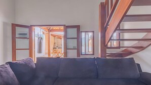 A living room with a dark-colored sofa, a wooden staircase, and a view of an outdoor area with a wooden structure. - Casa em Ubatuba com churrasqueira e varanda ampla (Ubatuba)