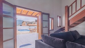 An open living space with a sofa, a staircase, and a view of a swimming pool and outdoor seating area. - Casa em Ubatuba com churrasqueira e varanda ampla (Ubatuba)
