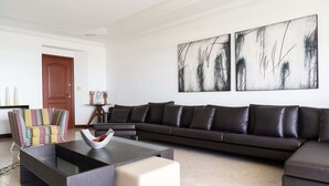 A modern living room with a sectional sofa, a colorful armchair, and a coffee table. There are abstract paintings on the wall and a wooden door in the background. - Apto em Cabo Frio em Frente a Praia com Varanda (Cabo Frio)