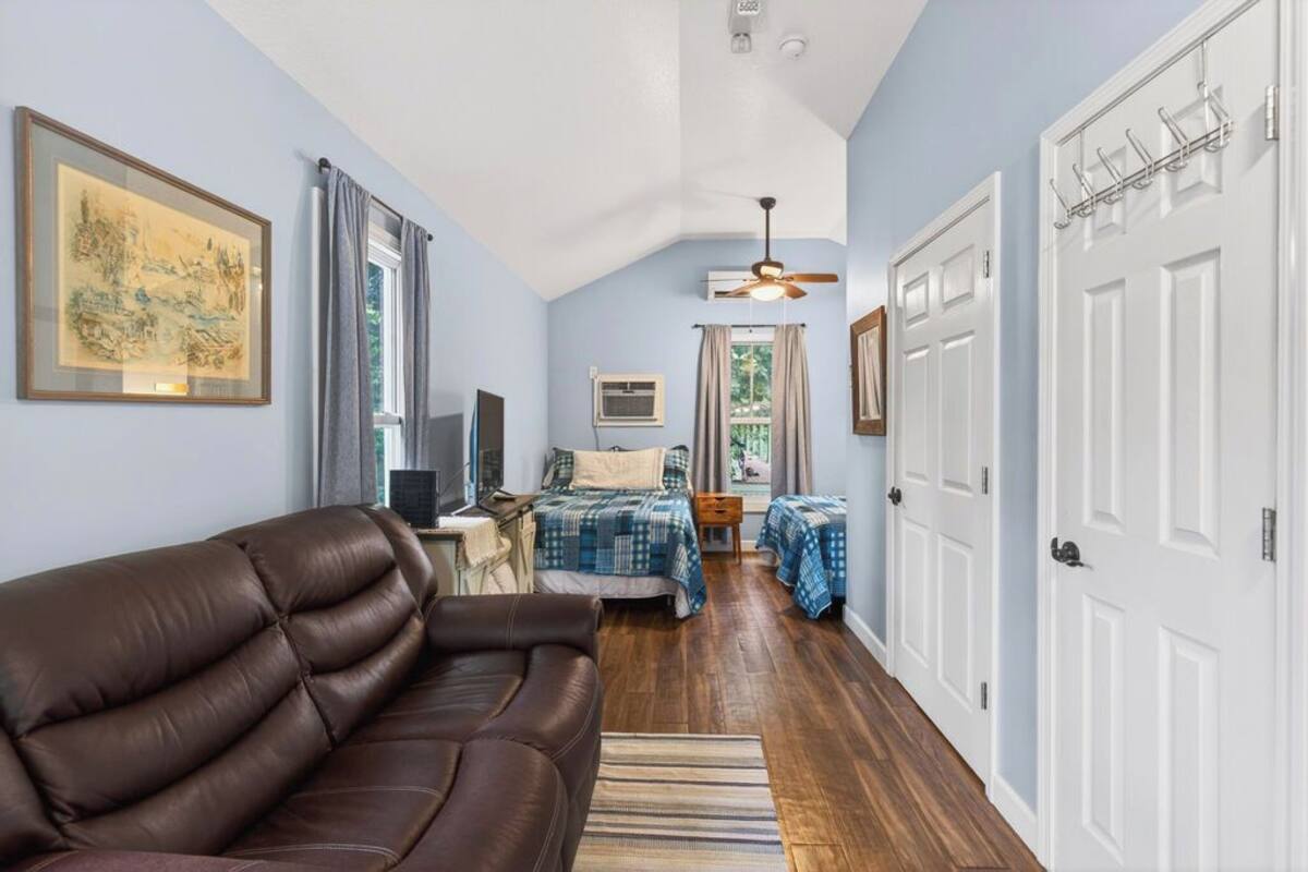 A room with a brown leather sofa, a bed with a blue checkered bedspread, a ceiling fan, and a hallway with white doors.