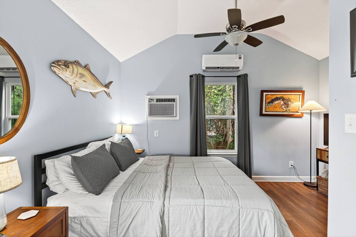 A bedroom with a bed, ceiling fan, air conditioning unit, window with curtains, and a fish decoration on the wall.