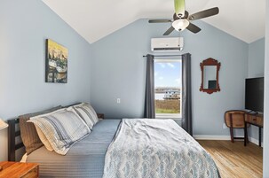 A bedroom with a bed, a desk, a chair, a ceiling fan, an air conditioner, a window with a view, and a painting on the wall.