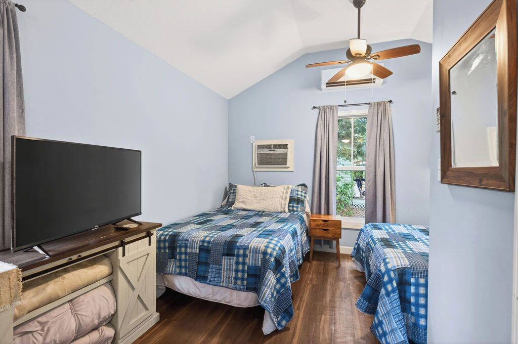 Een slaapkamer met een flatscreen televisie, twee bedden met geruit beddengoed, een plafondventilator, een airconditioning en een raam met gordijnen.