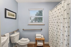 Un baño con inodoro, una mesita con toallas, una ventana con persianas y cortina de ducha.