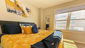 A bedroom with a large bed, a window with blinds, and artwork on the wall.