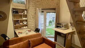 A cozy room with a brown sofa, a small dining table, a kitchen area with a microwave, and a view of the garden through a glass door. - Le Nid Charentais (Rouillac)