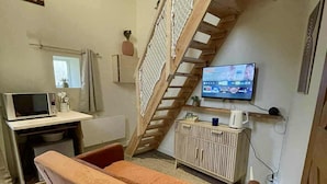 A cozy living room with a wooden staircase, a television, a sofa, and a small kitchen area. - Le Nid Charentais (Rouillac)