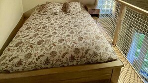 A bedroom with a wooden ceiling, a bed with floral bedding, a small table, and a window with a view of greenery. - Le Nid Charentais (Rouillac)