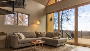 A modern living room with a sectional sofa, a small wooden coffee table, and a large window with a view of trees. - Highland Cows/Wood-Fired Hot Tub/Stunning Views (Logan)