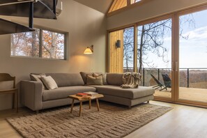 A modern living room with a sectional sofa, a small wooden coffee table, and a large window with a view of trees. - Highland Cows/Wood-Fired Hot Tub/Stunning Views (Logan)
