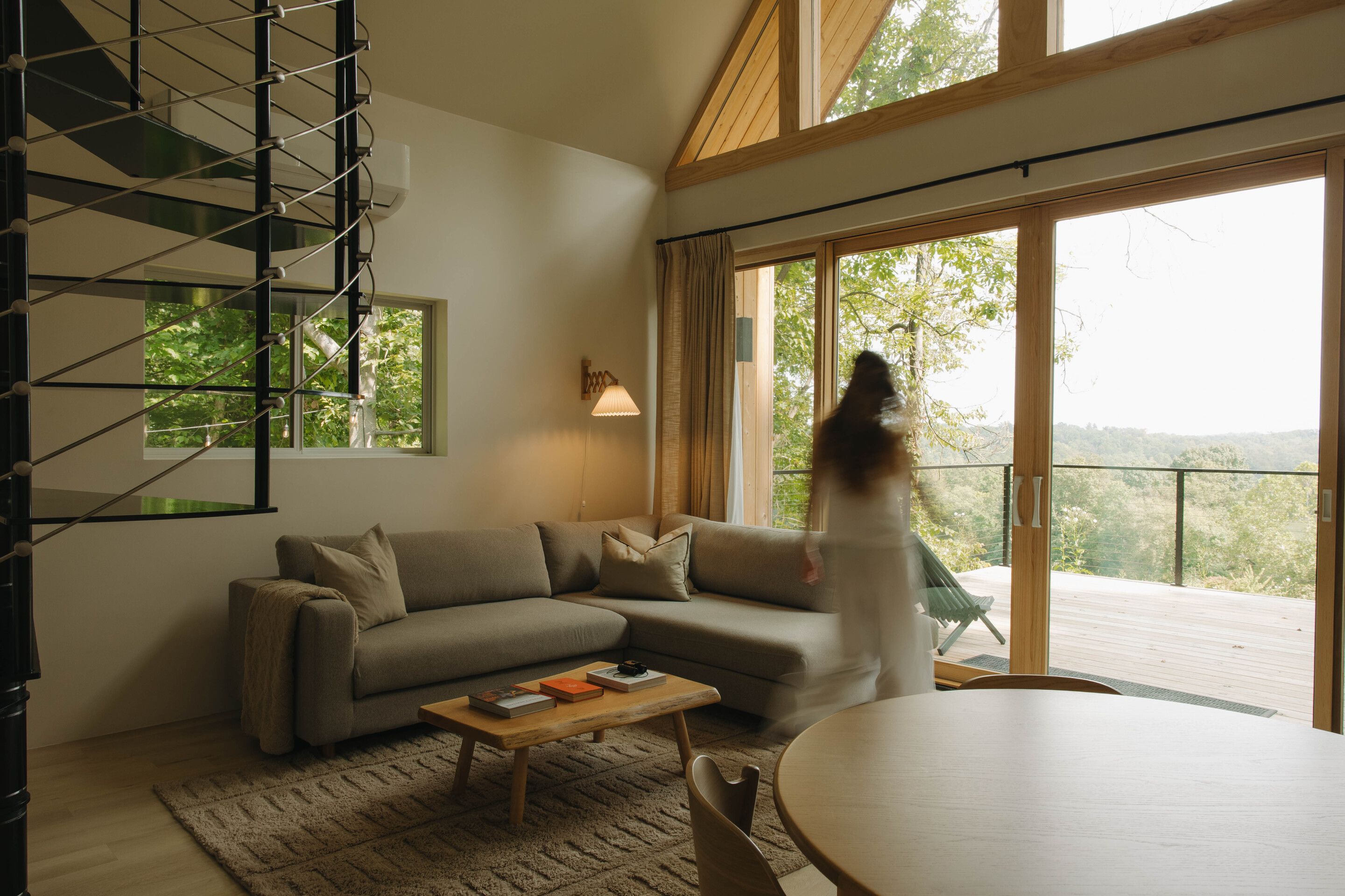A modern living room with a sectional sofa, a coffee table with books, a staircase, and a large sliding glass door leading to an outdoor deck.