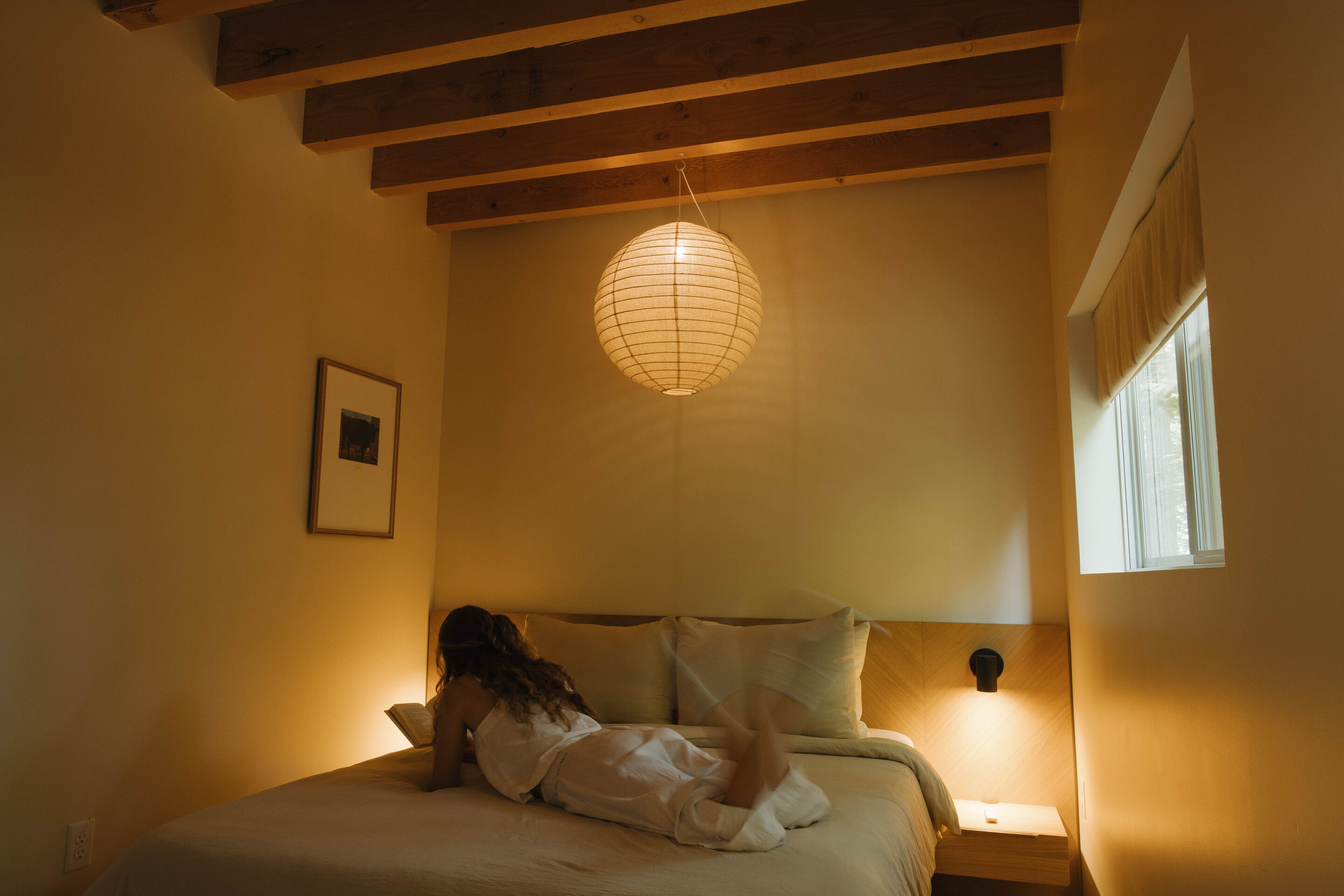 Una habitación con cama, una lámpara colgante, una ventana y un cuadro en la pared.