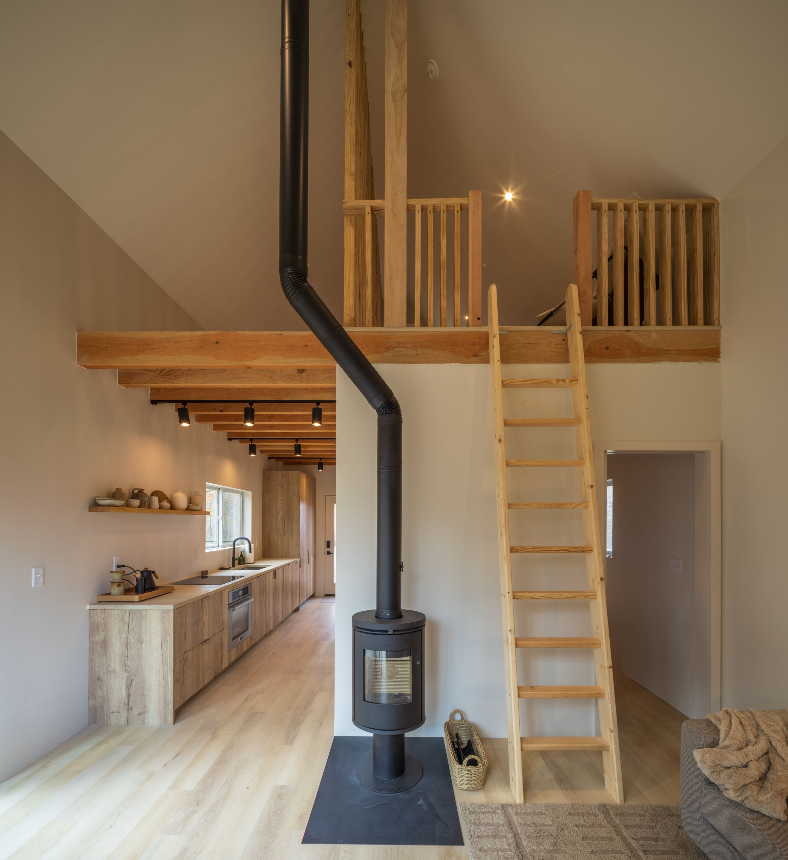 Un espacio habitable moderno estilo loft con vigas de madera expuestas, un estufa de leña negra en el centro, una escalera de madera y una cocina con armarios y encimera de madera.