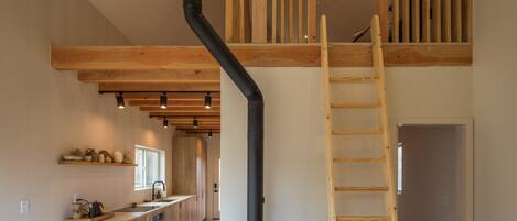 A modern loft-style living space with exposed wooden beams, a central black wood stove, a wooden ladder, and a kitchen area with wooden cabinets and a countertop.