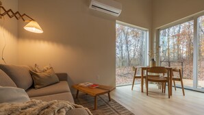 Un salon moderne avec un canapé, une table basse et un coin repas avec des chaises. Une grande fenêtre offre une vue sur les arbres.
