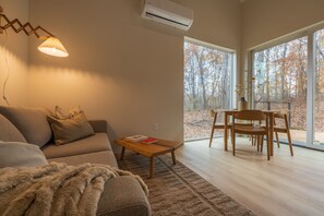 A modern living room with a sofa, a coffee table, and a dining area with chairs. There is a large window providing a view of trees. - Highland Cows/Wood-Fired Hot Tub/Forest Escape (Logan)