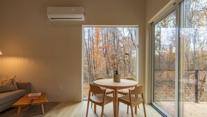 Un salon moderne avec une table ronde en bois et des chaises, un canapé et une grande fenêtre donnant sur la forêt.