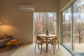 A modern living room with a round wooden table and chairs, a sofa, and a large window offering a view of trees. - Highland Cows/Wood-Fired Hot Tub/Forest Escape (Logan)