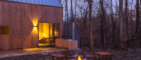 Una cabaña de madera con puerta de vidrio, un brasero y asientos al aire libre.