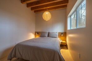 A bedroom with a large bed, wooden beams, a hanging light fixture, and a window with a view of trees. - Highland Cows/Wood-Fired Hot Tub/Forest Escape (Logan)