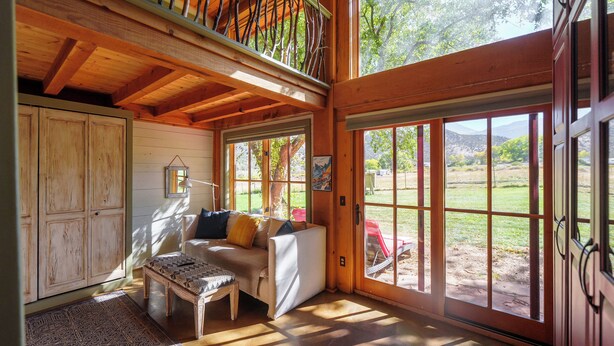 A cozy room with a wooden ceiling, a sofa, a patterned rug, and large windows offering a view of the outdoors.