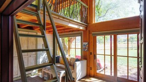 A spacious room with a high ceiling, a wooden staircase, a large window, and a door leading outside.