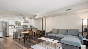 Een moderne woonkamer met een grijze hoekbank, een eethoek met een houten tafel en stoelen, en een keuken met witte kasten en een roestvrijstalen koelkast.