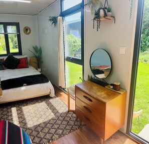A bedroom with a bed, a dresser, a mirror, and a window with a view of a garden. - 🌿 Coastal Cabin Retreat — Walk to Bethells Beach, Café & Freshwater Lake 🌞 (Te Henga (Bethells Beach))
