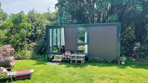 A small, modern outdoor structure with a large window, surrounded by lush greenery and a grassy lawn. - 🌿 Coastal Cabin Retreat — Walk to Bethells Beach, Café & Freshwater Lake 🌞 (Te Henga (Bethells Beach))