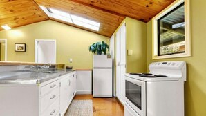 A compact kitchen with white appliances, a wooden ceiling, and a small window. - 🌿 Coastal Cabin Retreat — Walk to Bethells Beach, Café & Freshwater Lake 🌞 (Te Henga (Bethells Beach))