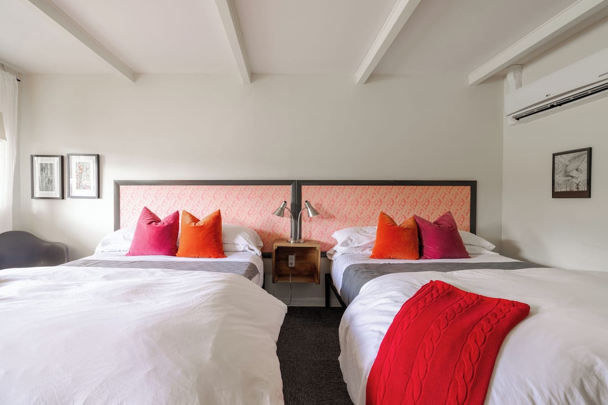 A hotel room with two beds, each with white bedding and colorful pillows. There is a wooden nightstand with a lamp between the beds. The room has white walls and a patterned wallpaper on the back wall.
