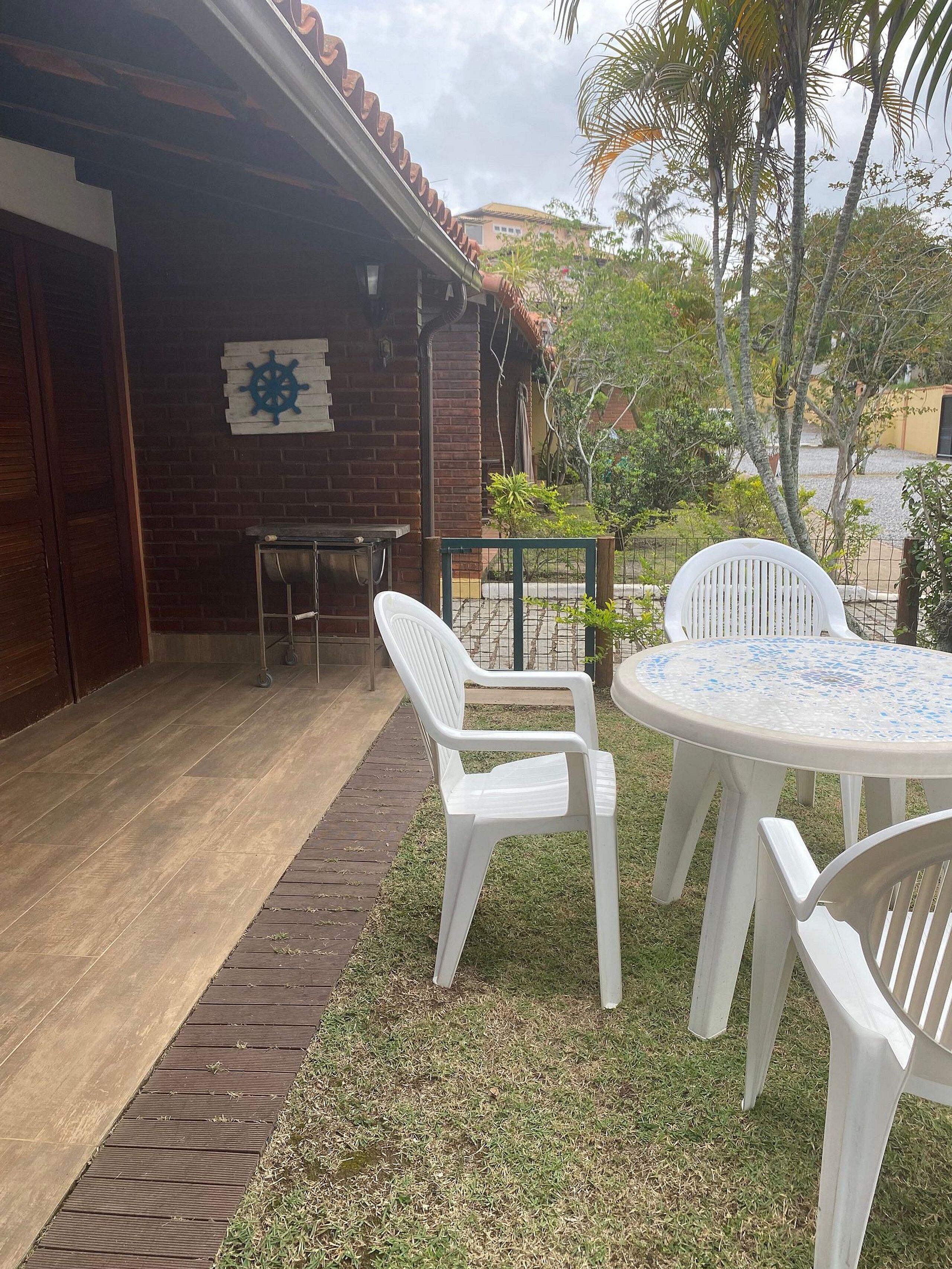 A patio area with a brick wall, a white plastic chair, a round table with a blue and white patterned top, and a small ship wheel decoration on the wall.