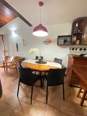 A dining area with a wooden table, black chairs, a hanging red pendant light, a white wall with a fish decoration, and a brick oven. - CASA DE PRAIA EM GERIBÁ (Búzios)