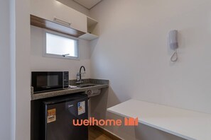 A compact kitchen with a microwave, sink, and refrigerator. There's a small window above the microwave and a white shelf above the sink. - Apto na Bela Vista ao lado da Nove de Julho (São Paulo)