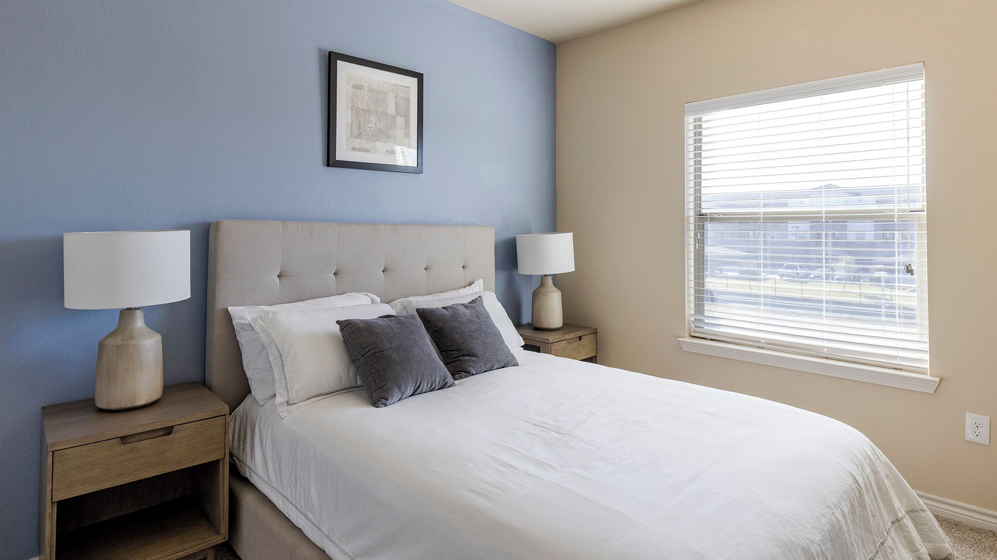 A bedroom with a bed, two bedside tables, two lamps, a framed picture on the wall, and a window with blinds.