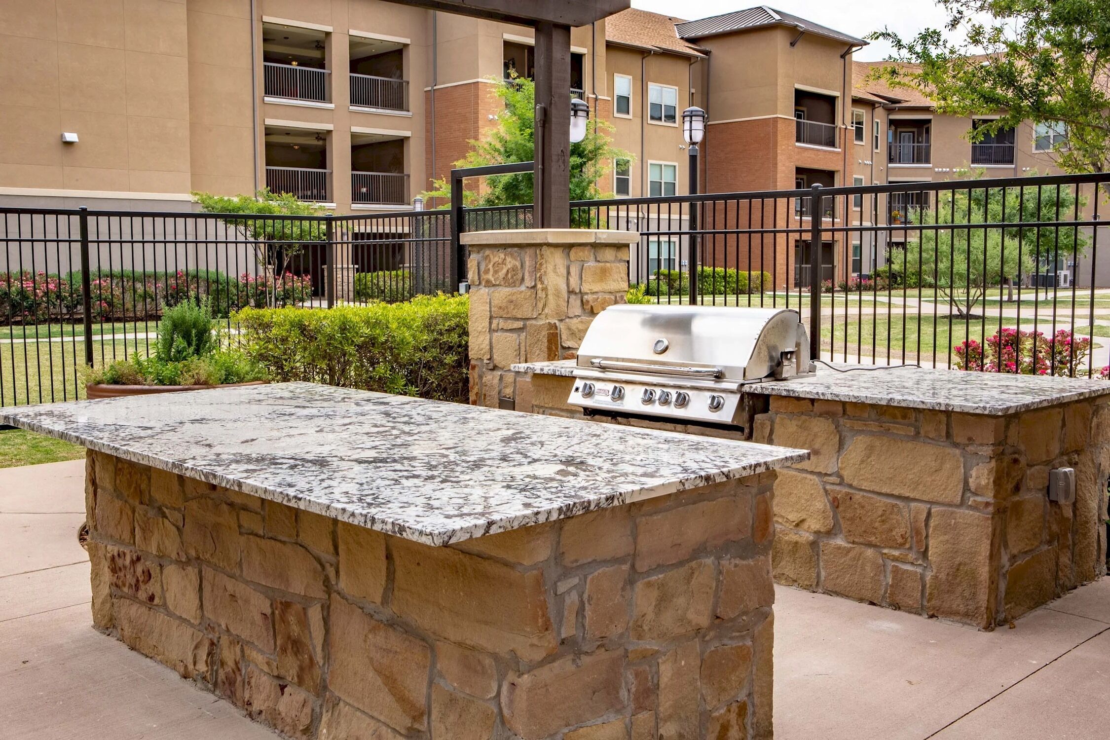 An outdoor barbecue area with a stone grill and a stone countertop, surrounded by a fence and greenery.