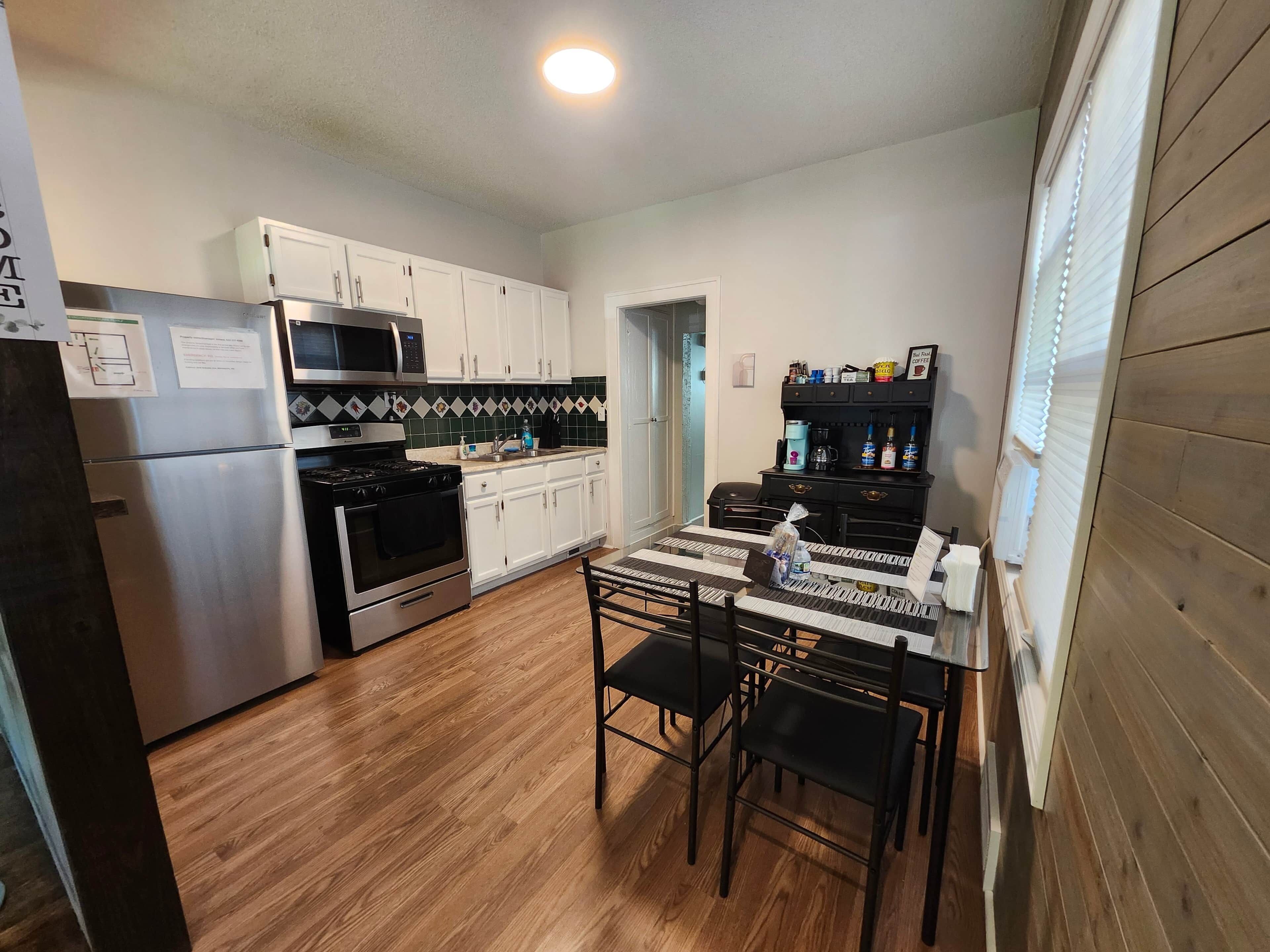 A compact kitchen with white cabinets, a stainless steel refrigerator, a microwave, and a stove. There is a dining table with black chairs and a coffee station with a coffee maker and bottles.