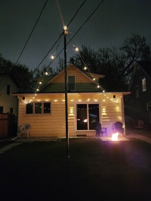 Una casa de un piso con porche, una fogata y luces colgantes.