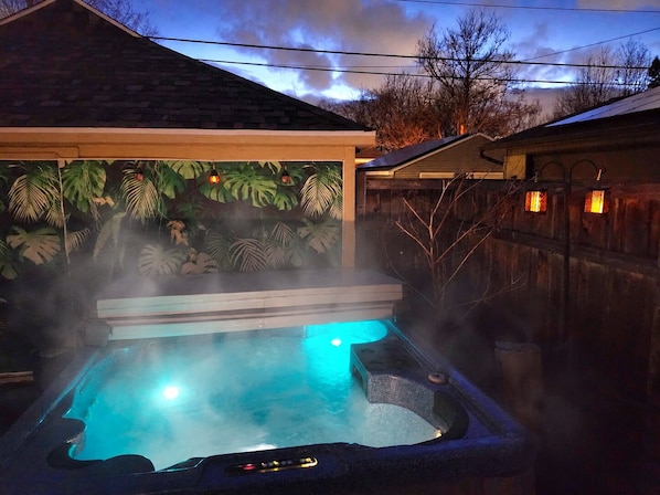 A hot tub with steam rising, surrounded by a wooden fence and a backdrop of tropical foliage.
