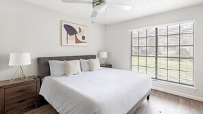 A bedroom with a bed, two bedside tables, a ceiling fan, a window with blinds, and a framed artwork on the wall. - Landing Apartments near Southridge (Denton)