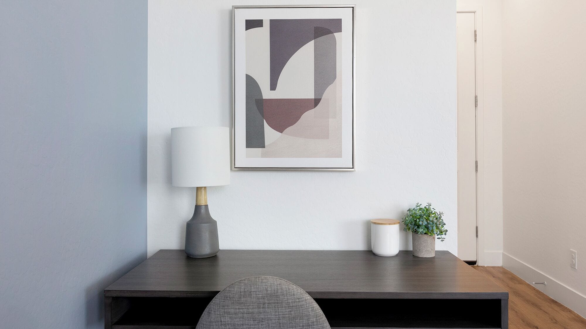 A modern, minimalist room with a dark wood desk, a grey chair, a white lamp, a framed abstract artwork, and a small potted plant.