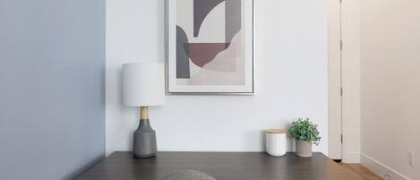 A modern, minimalist room with a dark wood desk, a grey chair, a white lamp, a framed abstract artwork, and a small potted plant.
