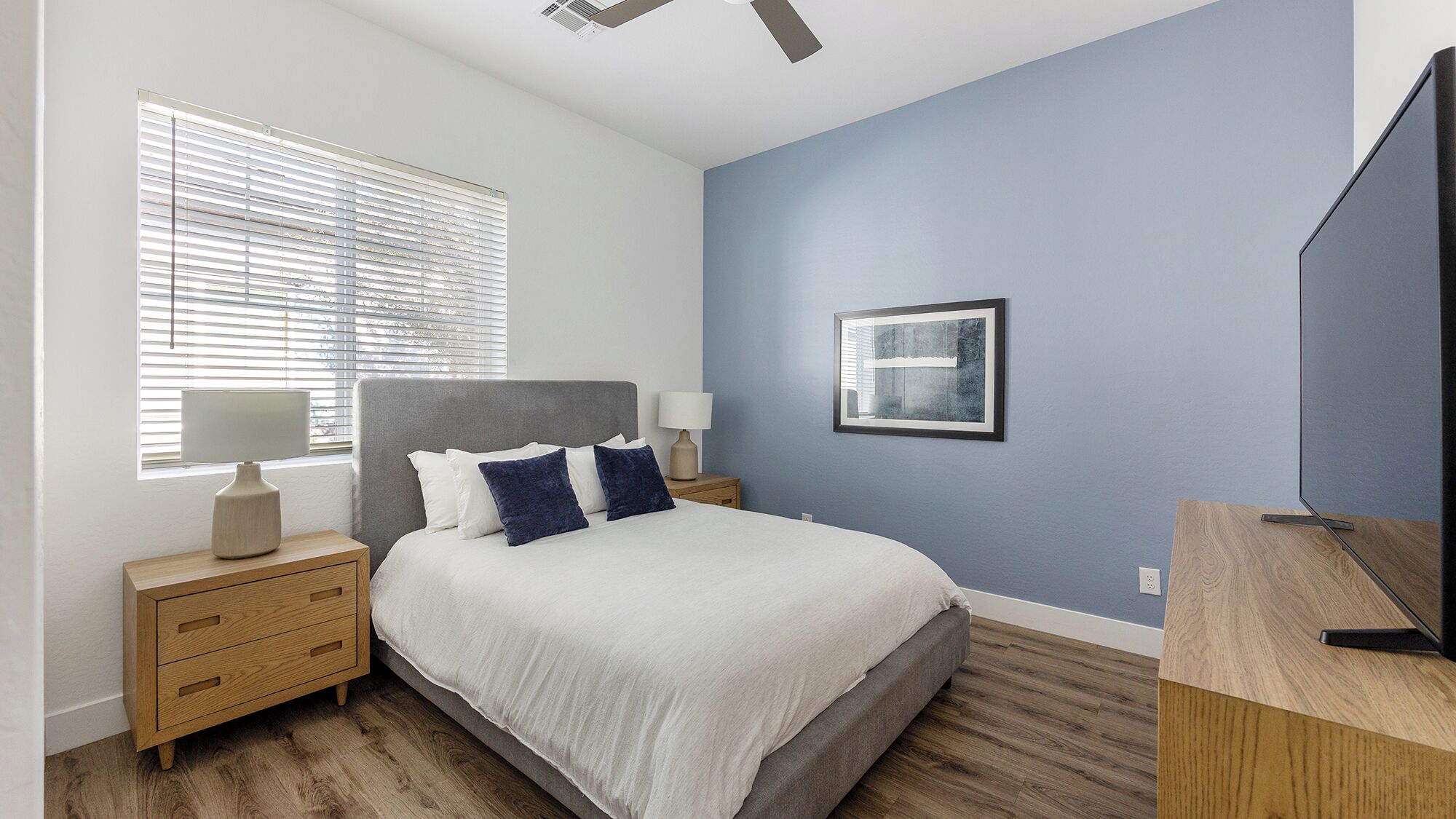 A bedroom with a bed, bedside tables, a TV, and a window with blinds.