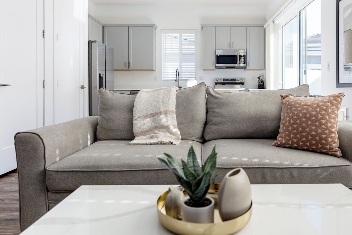 Una sala de estar moderna con un sofá gris, una mesa de centro con adornos, y una cocina al fondo.