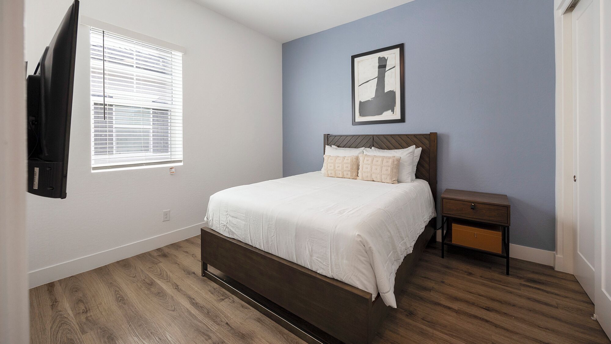 A bedroom with a bed, a nightstand, a wall-mounted TV, and a window with blinds.